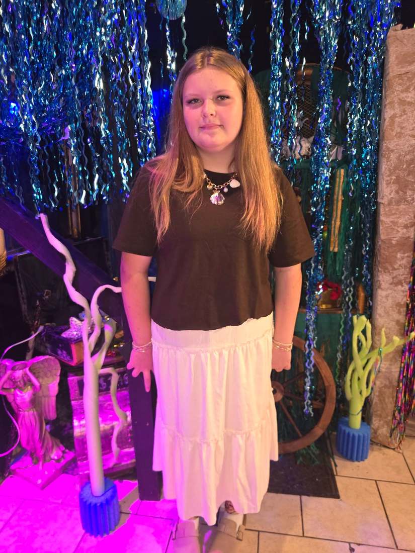 A Little Mermaid cast member in a brown top and white tiered skirt wearing a shell necklace