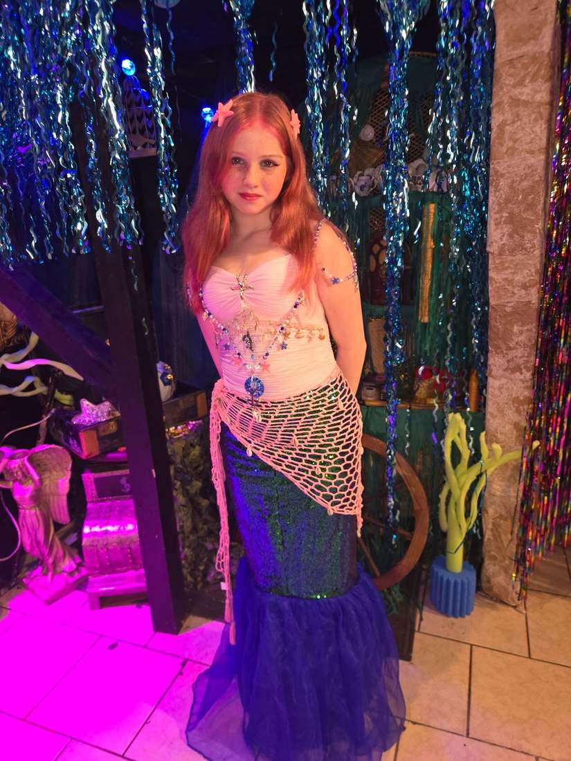 Ariel with red hair, a pink shell top, and a green and blue mermaid tail draped with netting