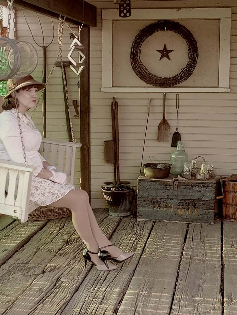 A cast member in a white 1930s dress and hat sitting on a porch swing