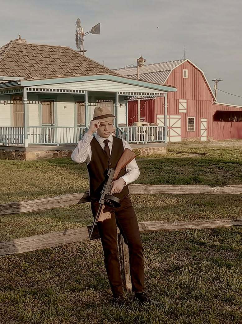 Clyde tipping his fedora and holding a Tommy gun by a fence, with a farmhouse and red barn behind him