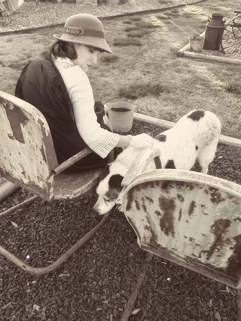 A cast member in a cloche hat petting a spotted dog beside rusted metal chairs (sepia)