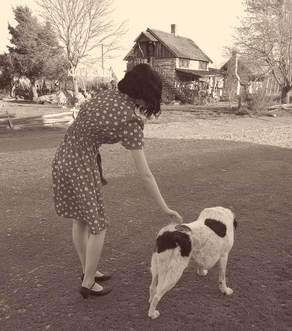 A cast member in a floral dress bending down to a spotted dog outside a rustic cabin (sepia)