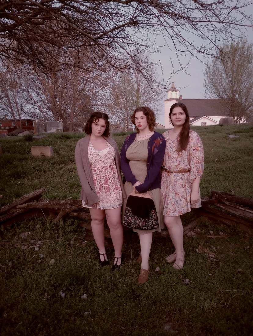 Three cast members in period dresses standing on a grassy hill with a white church behind them