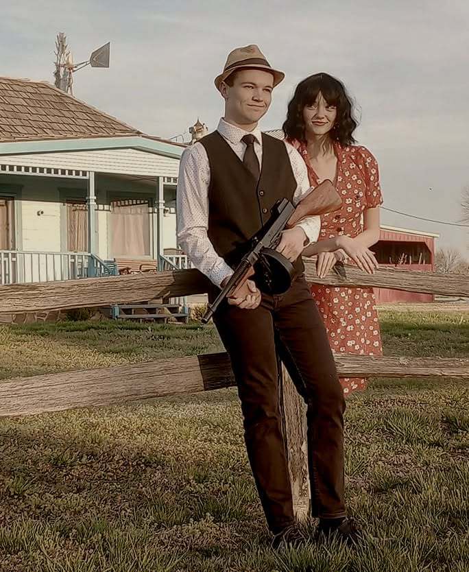 Two cast members leaning on a split-rail fence in front of the farmhouse