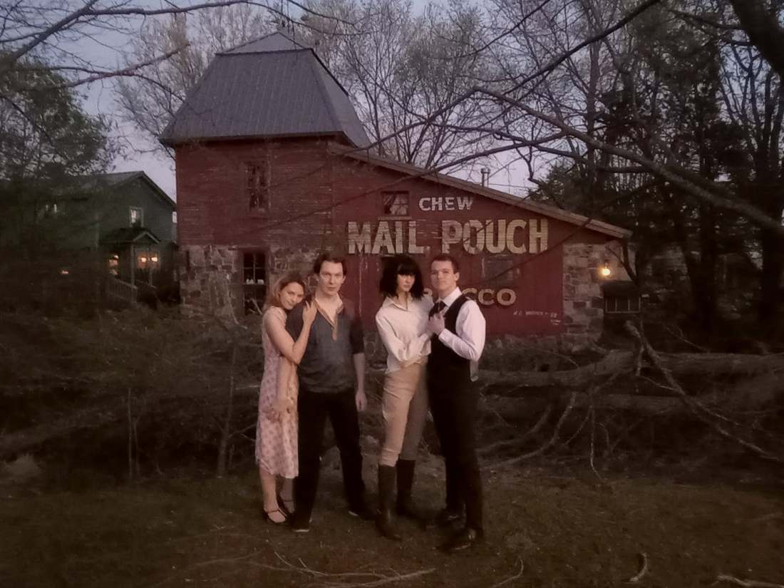 Four cast members in front of a red barn painted with a Chew Mail Pouch Tobacco sign at dusk