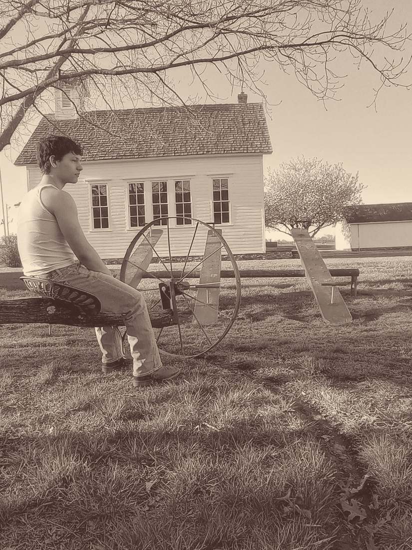 A cast member in a tank top sitting on a merry-go-round in front of a white schoolhouse (sepia)