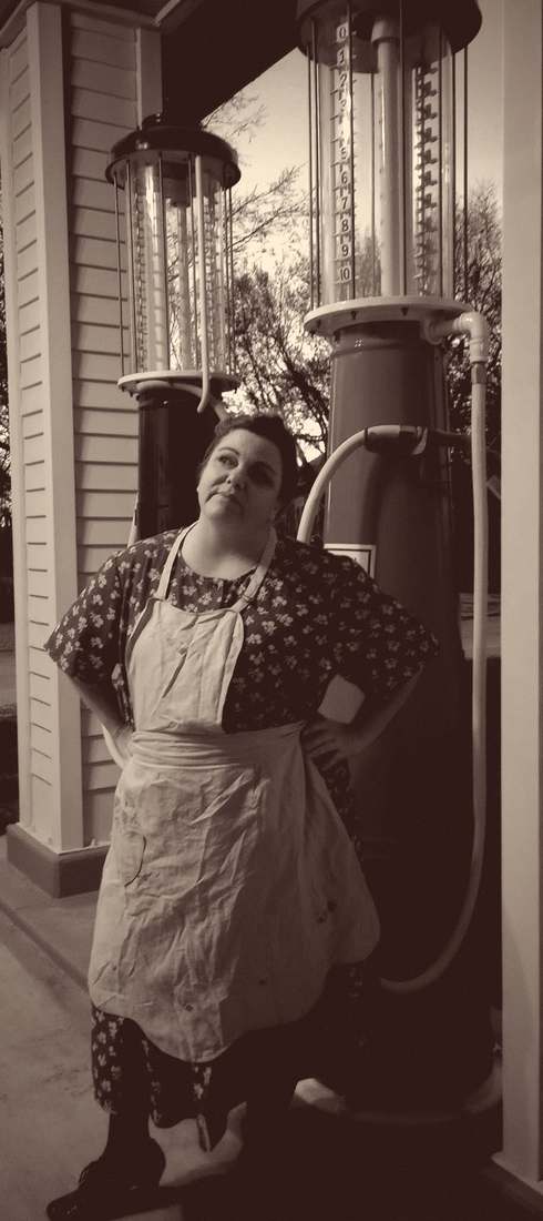 A cast member in a floral dress and apron leaning against vintage glass-cylinder gas pumps (sepia)