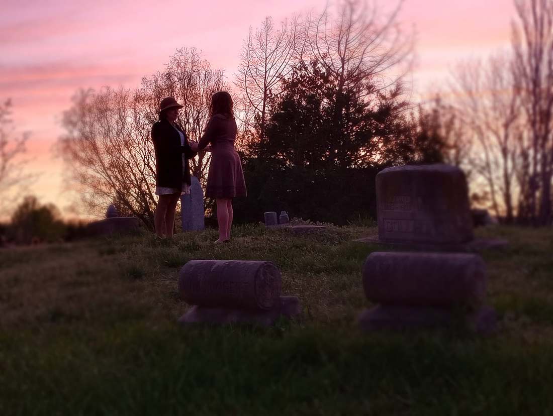 Two cast members holding hands among gravestones at sunset