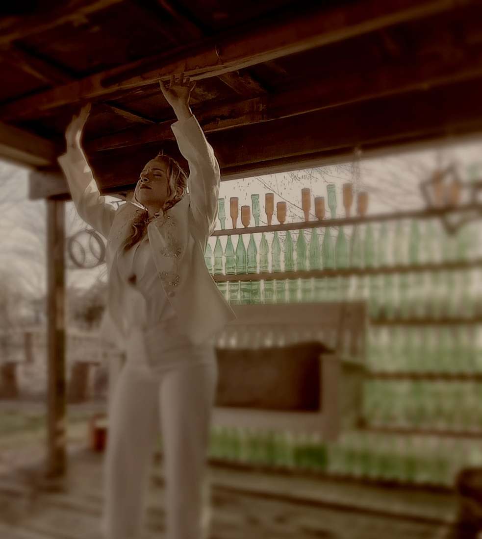 A cast member in a white suit reaching up to a porch beam, a wall of green bottles behind her (sepia)
