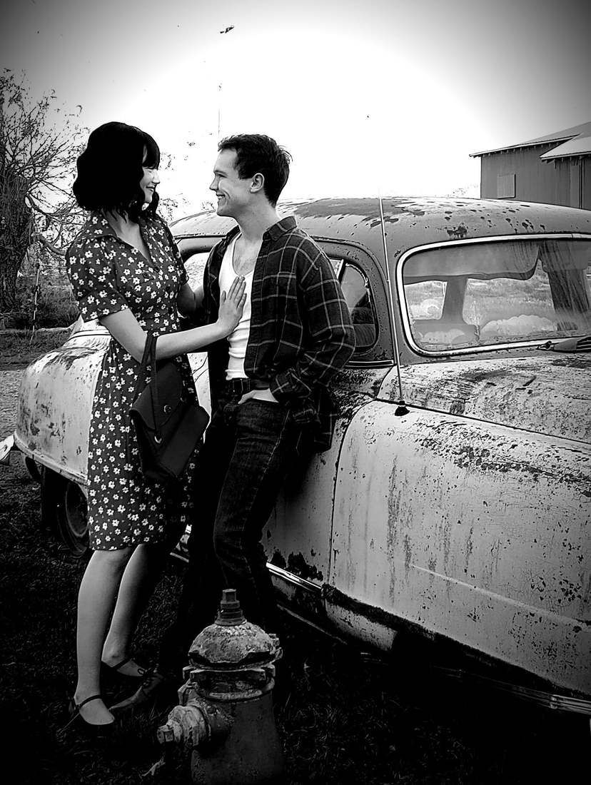 Bonnie and Clyde smiling at each other beside a rusted vintage car (black and white)