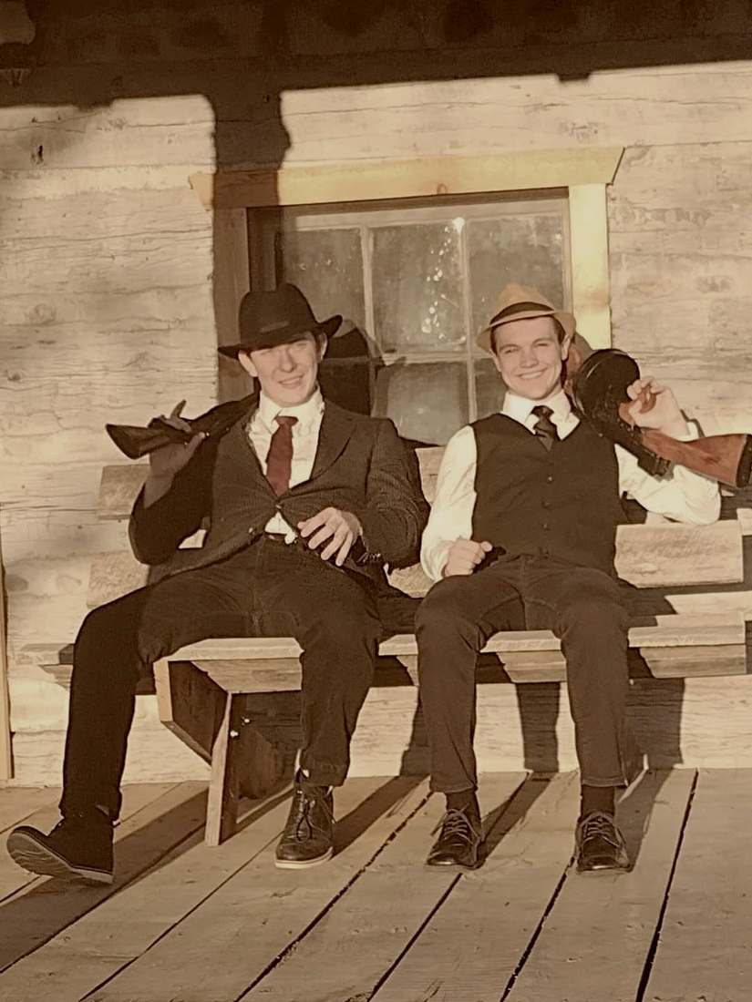 Two cast members with Tommy guns smiling on a cabin porch bench (sepia)