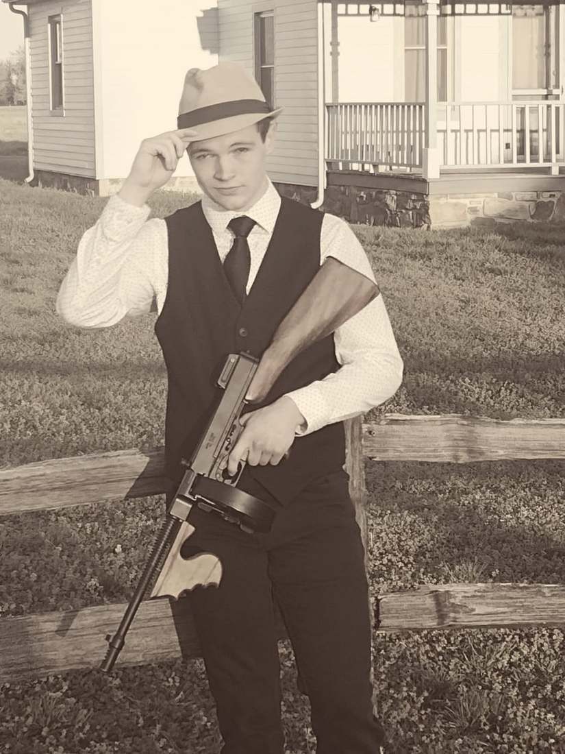 Clyde tipping his fedora and holding a Tommy gun by a split-rail fence (sepia)
