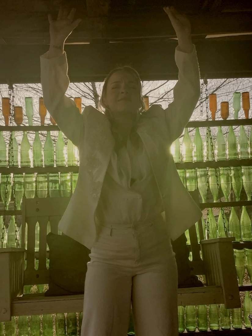A cast member in a white suit backlit by the green bottle wall, arms raised