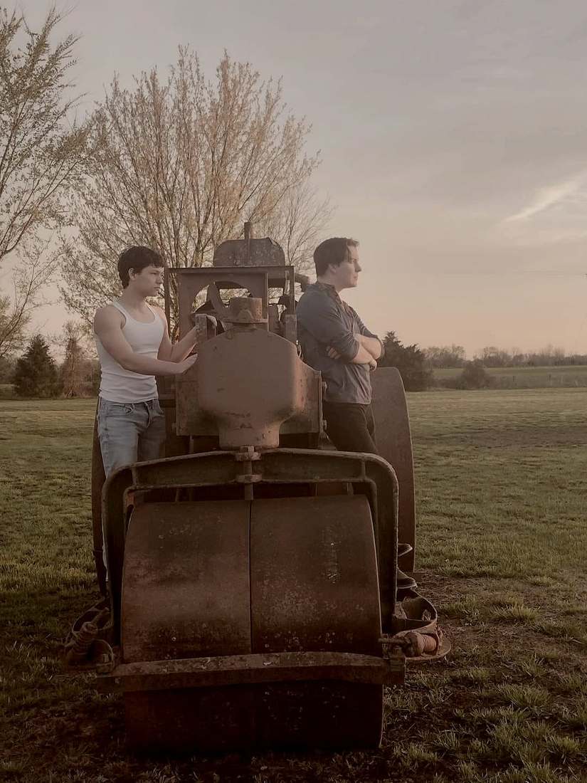 Two cast members standing on a vintage steamroller in a field at dusk