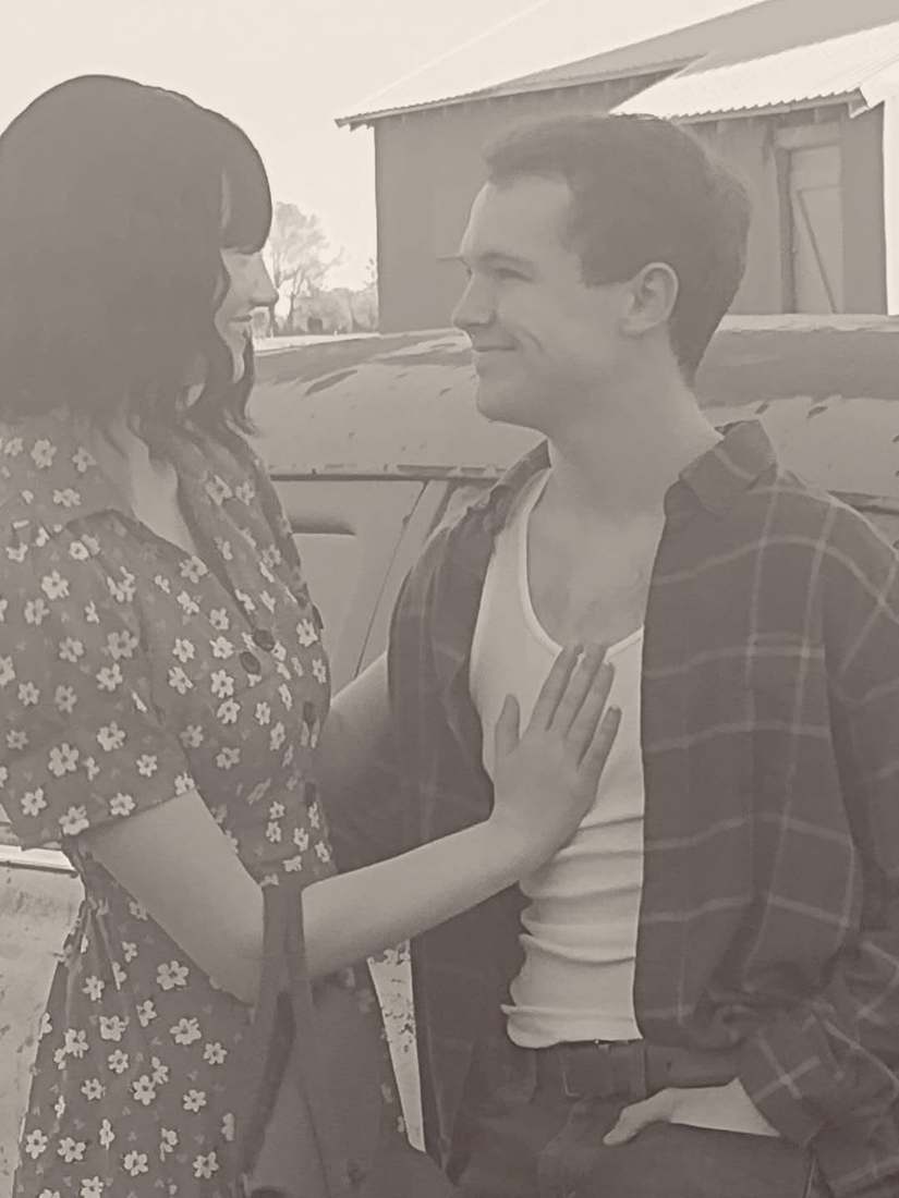 Bonnie and Clyde smiling at each other beside the vintage car, close up (sepia)