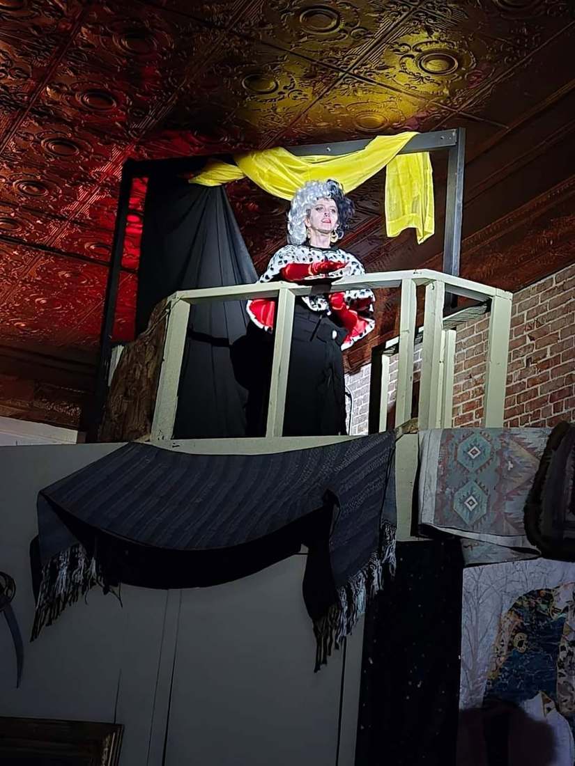 A grey-wigged performer in ruffles watches from the balcony railing in Twisted