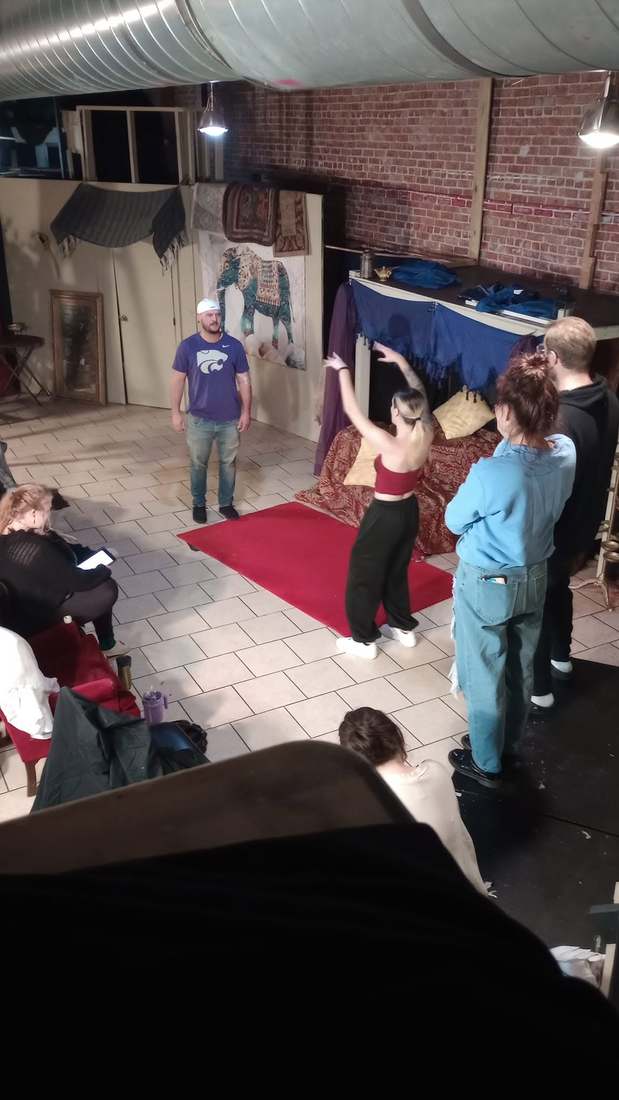 A performer gestures through a scene in rehearsal while castmates watch from the set