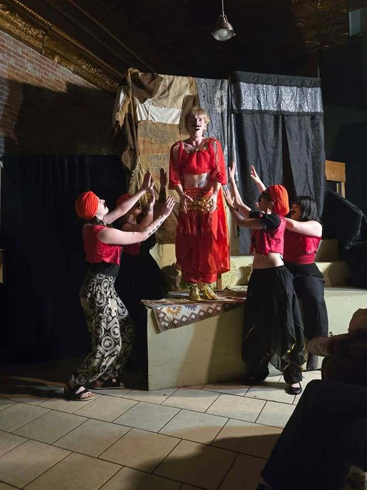 Dancers in red reach up toward a performer standing on the platform in Twisted