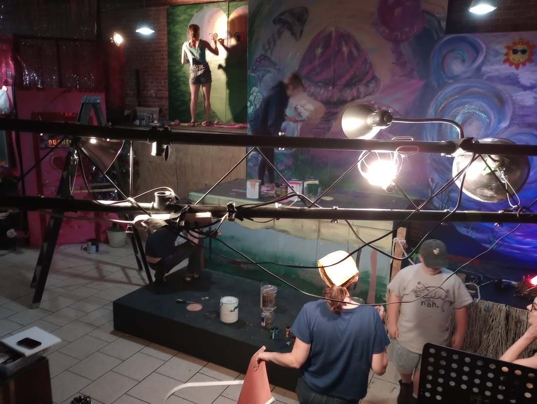 Company members painting a large colorful backdrop flat inside the studio, paint cans and work lights scattered around